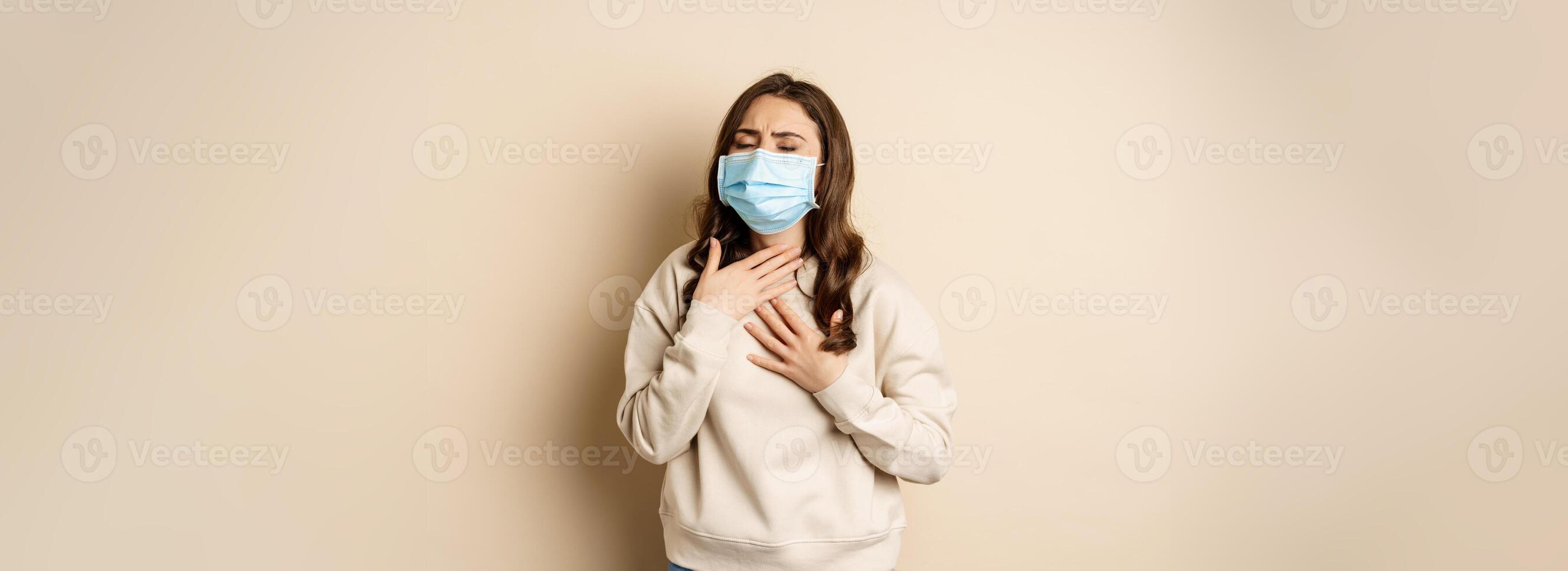 Covid-19 and health concept. Sick woman in medical face mask coughing, feeling ill with sour throat, standing over beige background photo