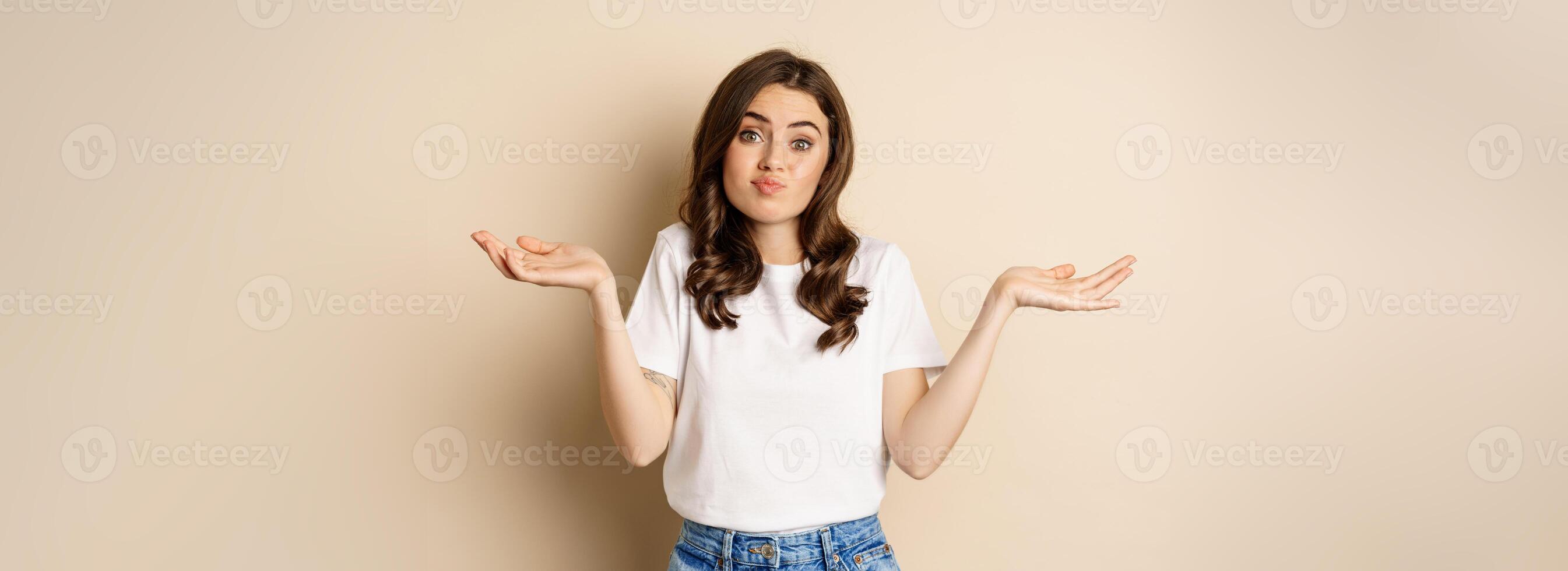 Young confused woman shrugging shoulders and with clueless face expression, standing over beige background photo