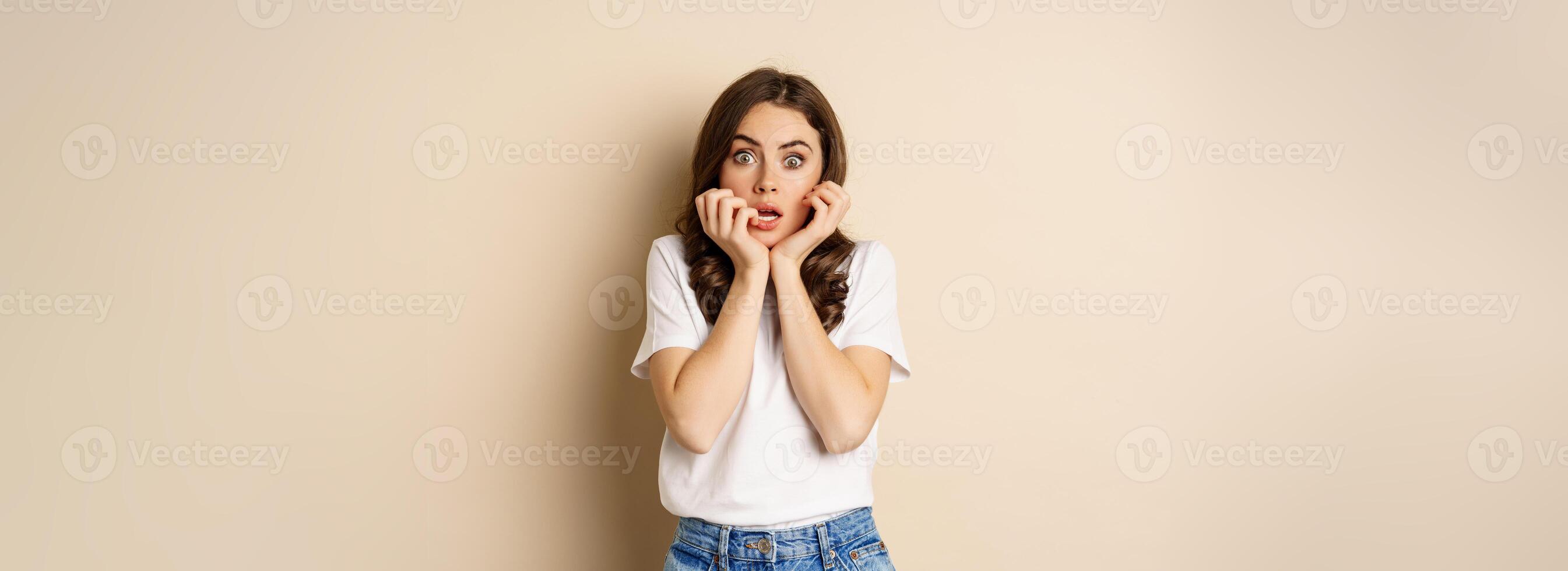 Scared and shocked woman gasping, looking startled speechless, wearing t-shirt, standing over beige background photo