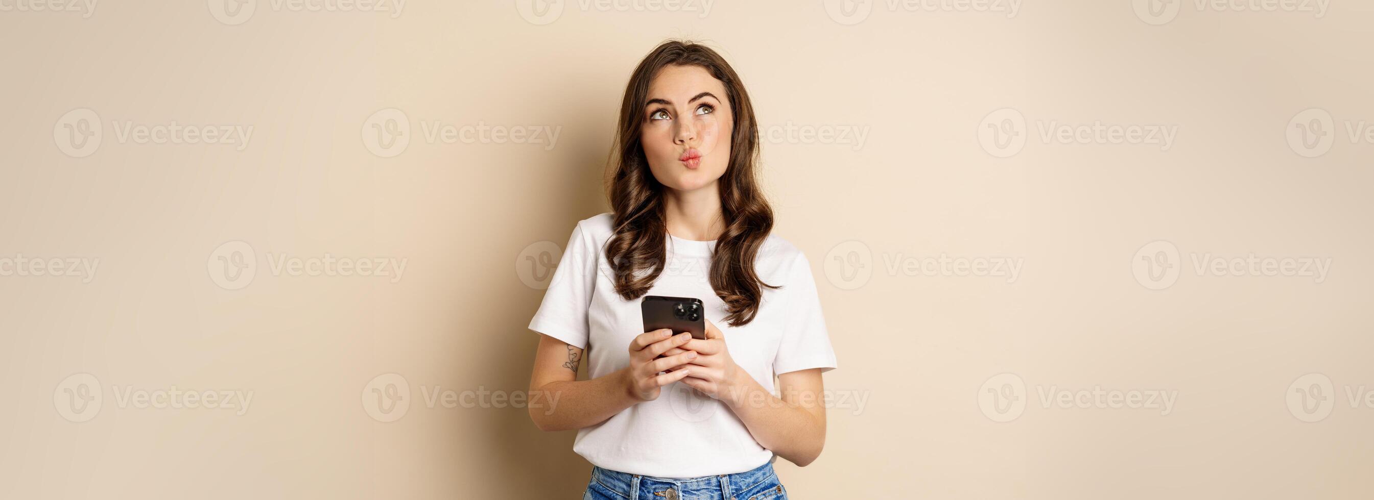 Portrait of woman thinking, holding mobile phone and looking up with thoughtful face, standing over beige background photo