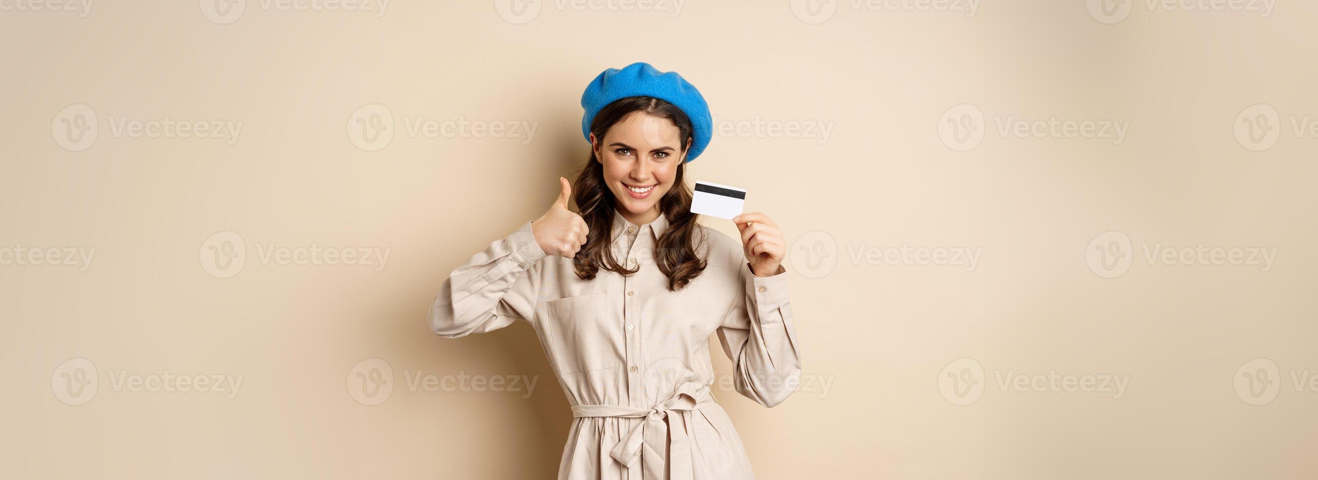 Concept of shopping and sale. Happy young woman in stylish clothes, showing credit card store discount and thumbs up, recommending, standing over beige background photo