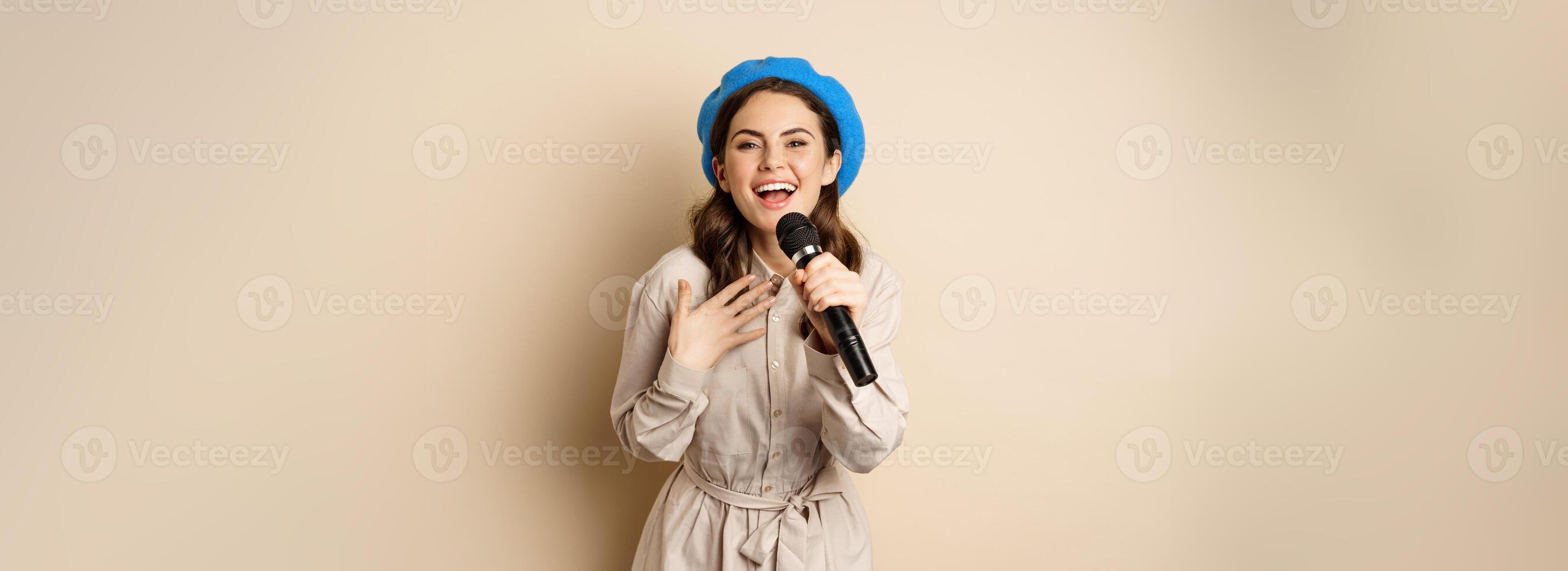 Portrait of happy stylish woman performing, singing with microphone, posing against beige background photo