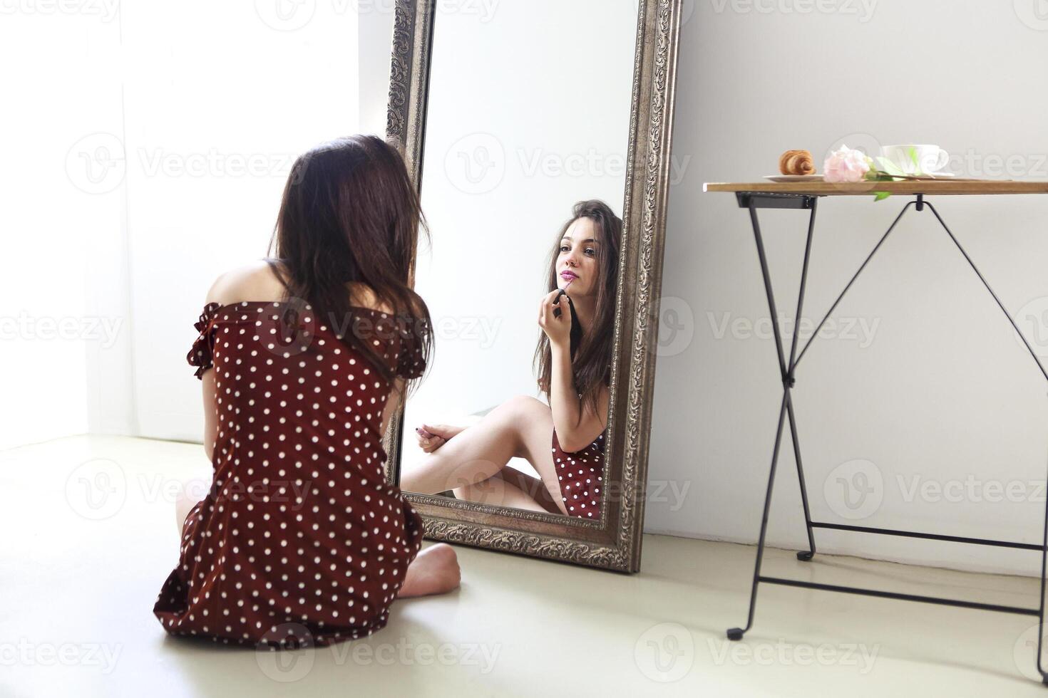 Beautiful woman in polka dots dress applying lipstick and looking at mirror photo