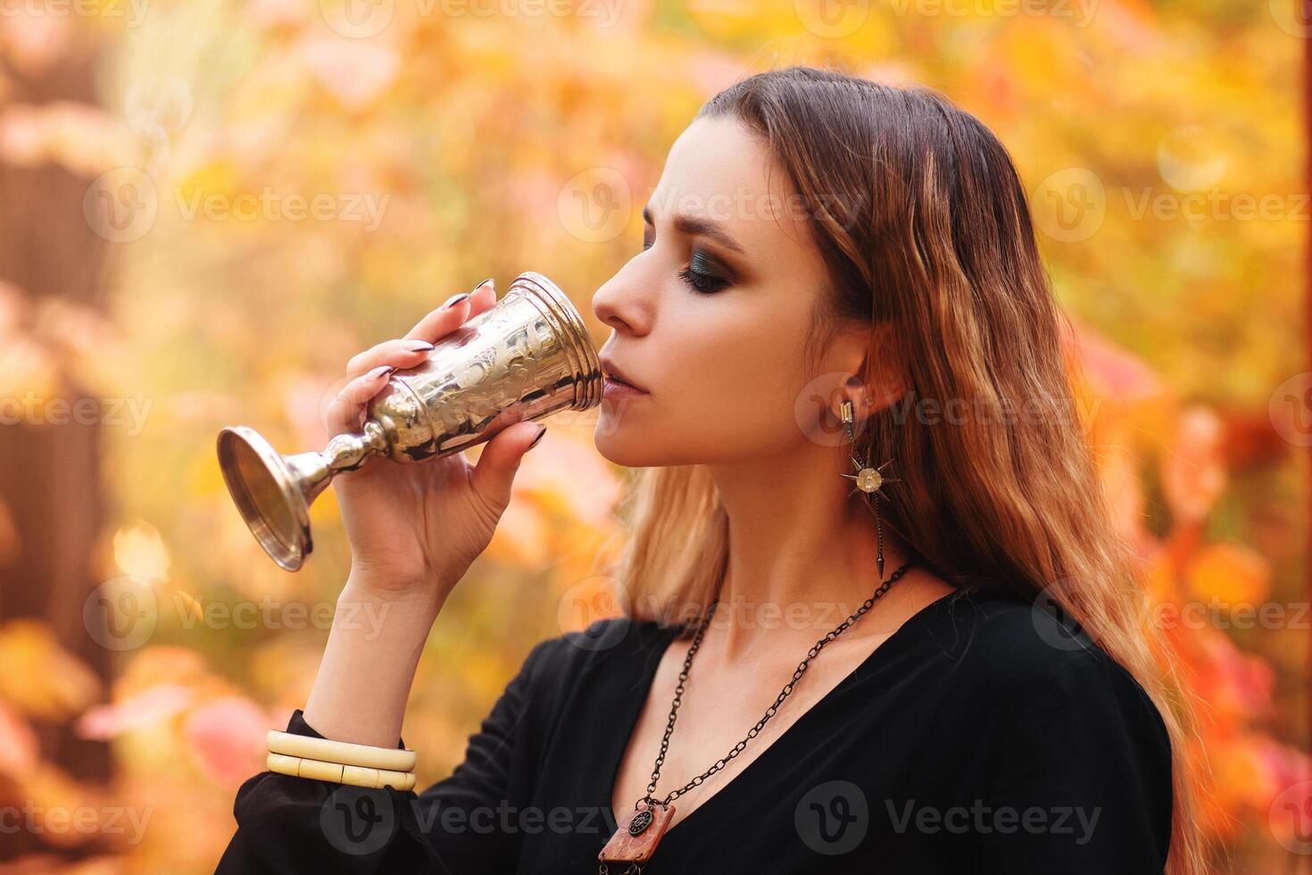Young cheerful woman in black dress holding vintage metal glass in hands while standing in autumn park photo