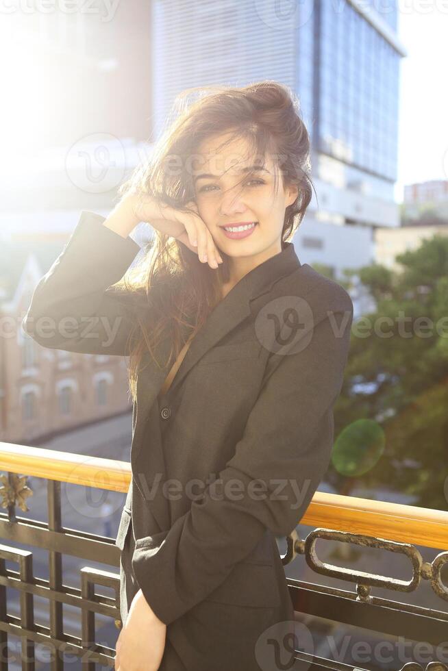 Young woman in jacket standing on balcony with city background at sunset photo