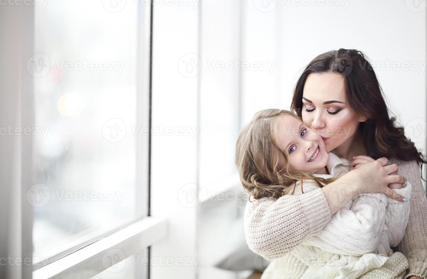 Mother and child daughter hugging by the window wearing cozy sweaters photo