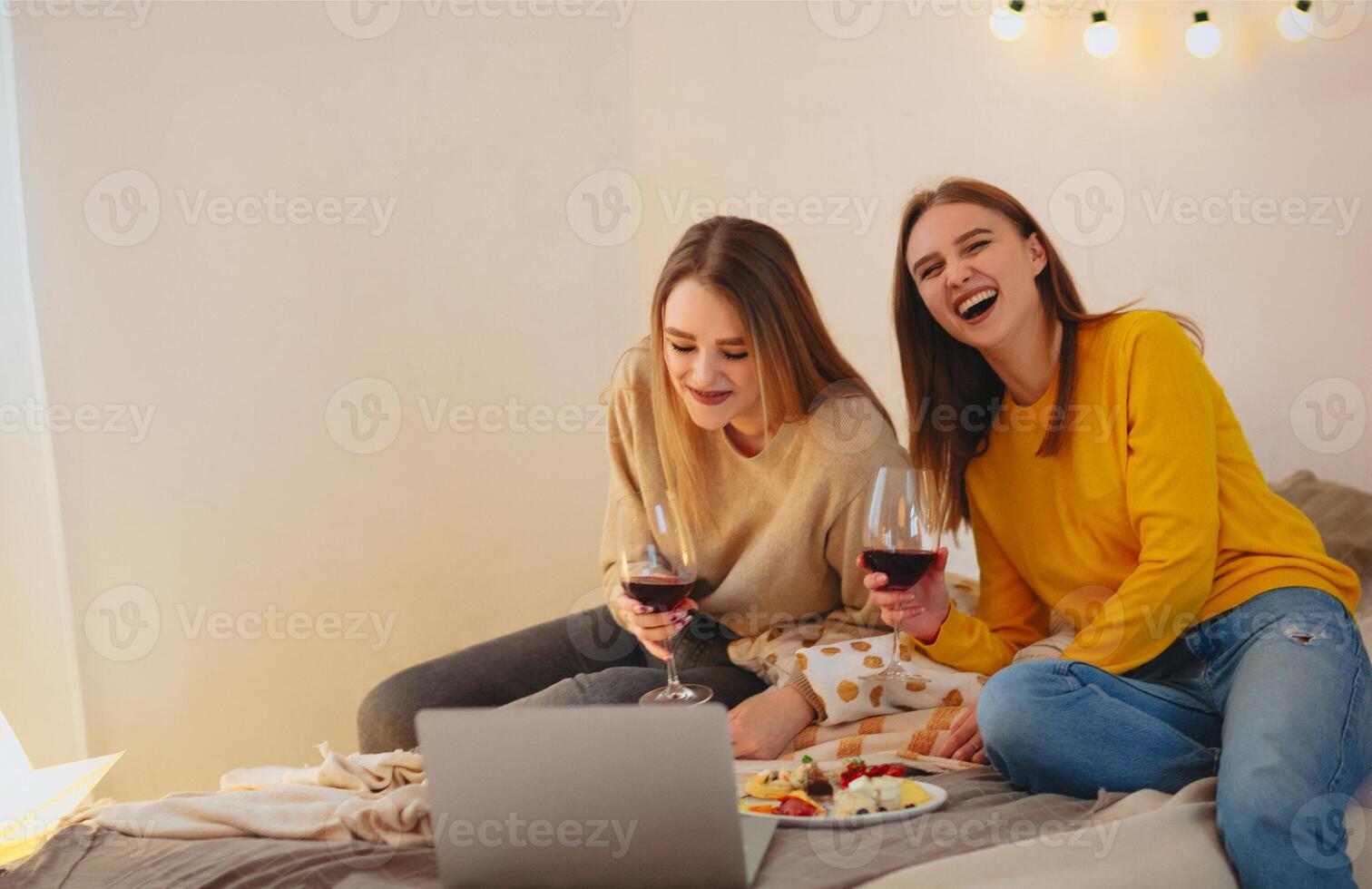 Two cute happy funny women having home cinema evening watching comedy on laptop sitting on bed photo