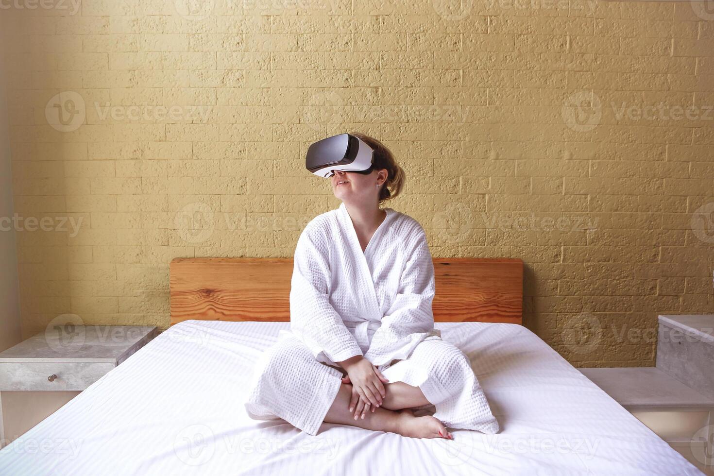 Amazed young woman sit wake up in white bed wearing virtual reality glasses, stunned female awaken rest in hotel or home bedroom try experience VR googles photo