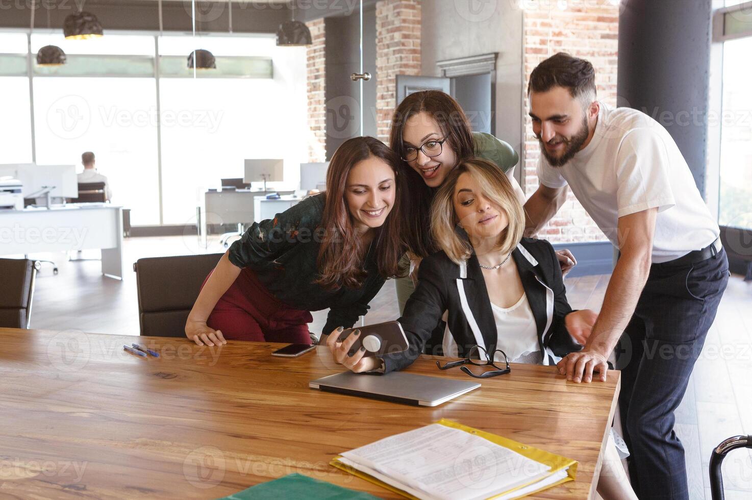 Smiling diverse colleagues gather in boardroom looking in mobile phone making selfi photo