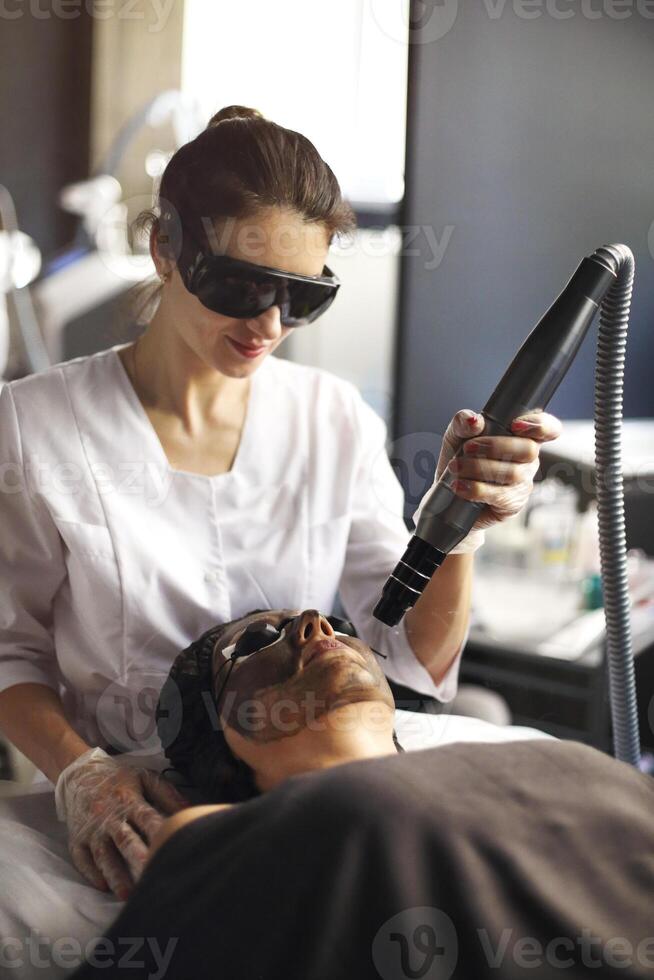 Beautician holding beauty laser near woman face covered with carbon mask and protective eyewear photo