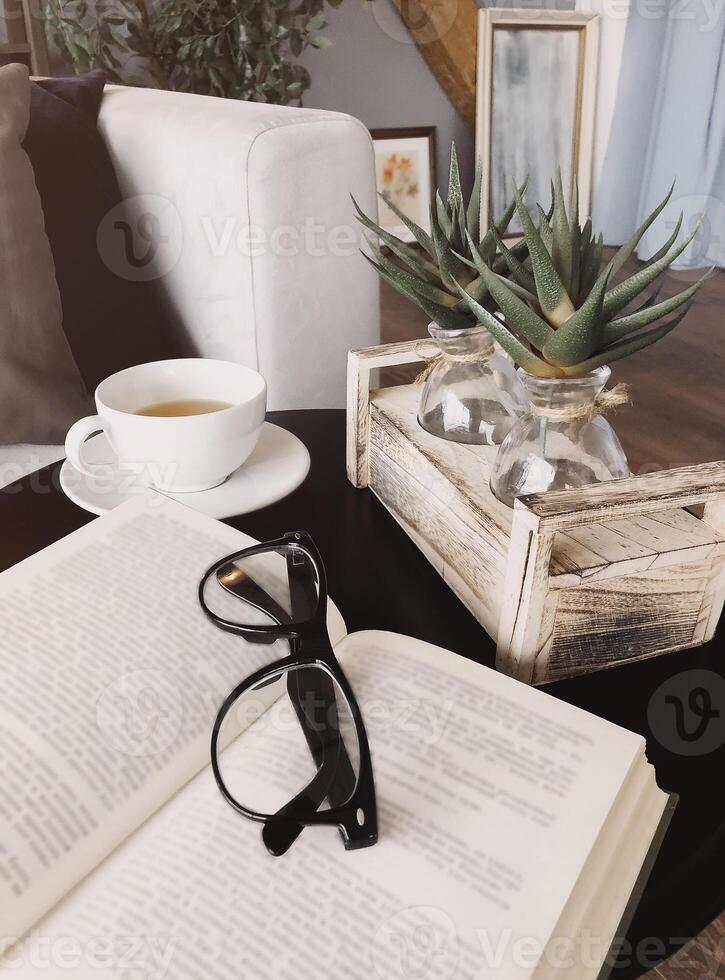 Eyeglasses on open book lying on round coffee table decorated with aloe plants photo
