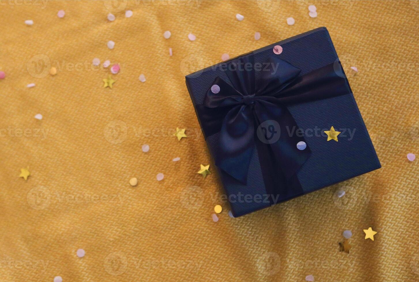 New Year or Christmas flat lay. Top view Xmas holiday celebration decorative black gift box with black bow, bright sparkles on the yellow background with copy space photo