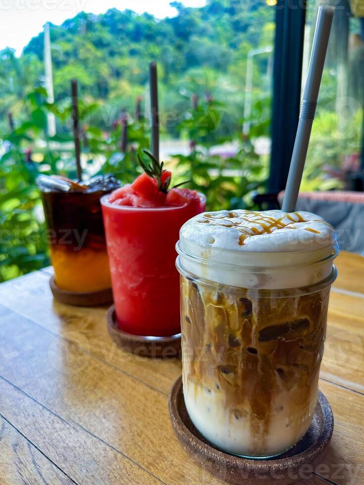 A selection of refreshing drinks including iced coffee, watermelon smoothie, and citrus iced tea on a wooden table, set against a lush tropical backdrop. Perfect for food and beverage themes. photo