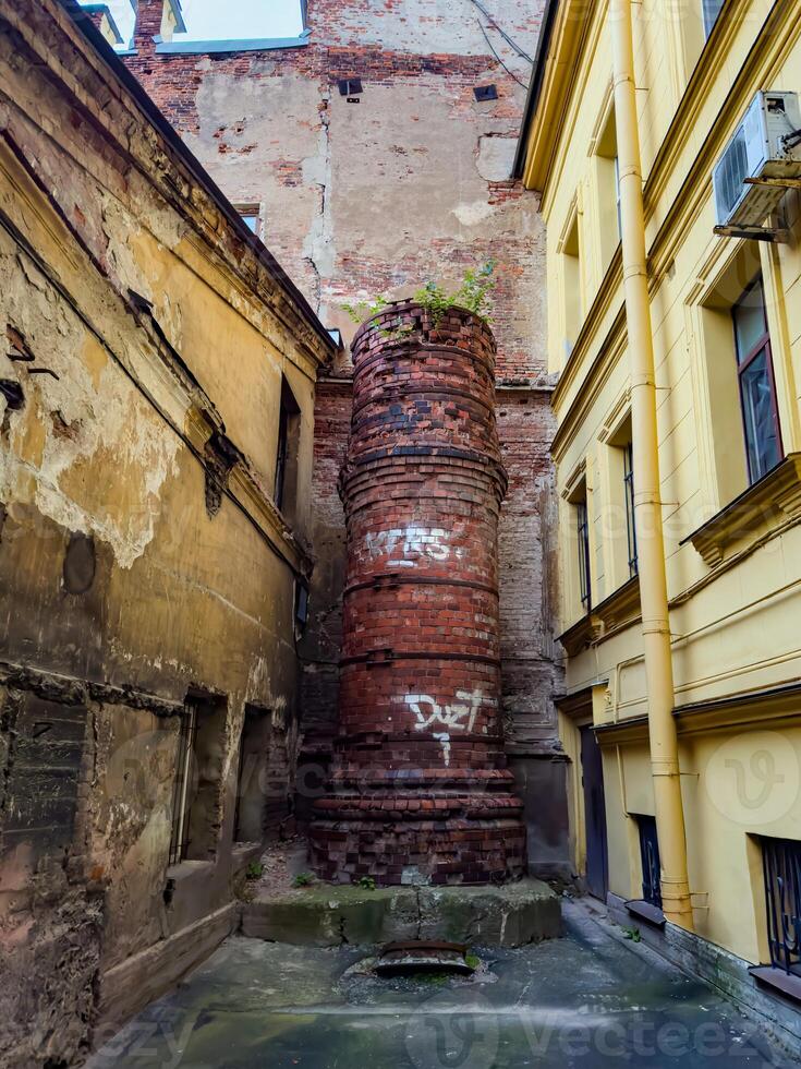 A view of an abandoned brick chimney in a dilapidated urban alley, surrounded by weathered walls and graffiti. The image captures the sense of decay and urban neglect, highlighting the textures and colors of the aged bricks and crumbling plaster photo