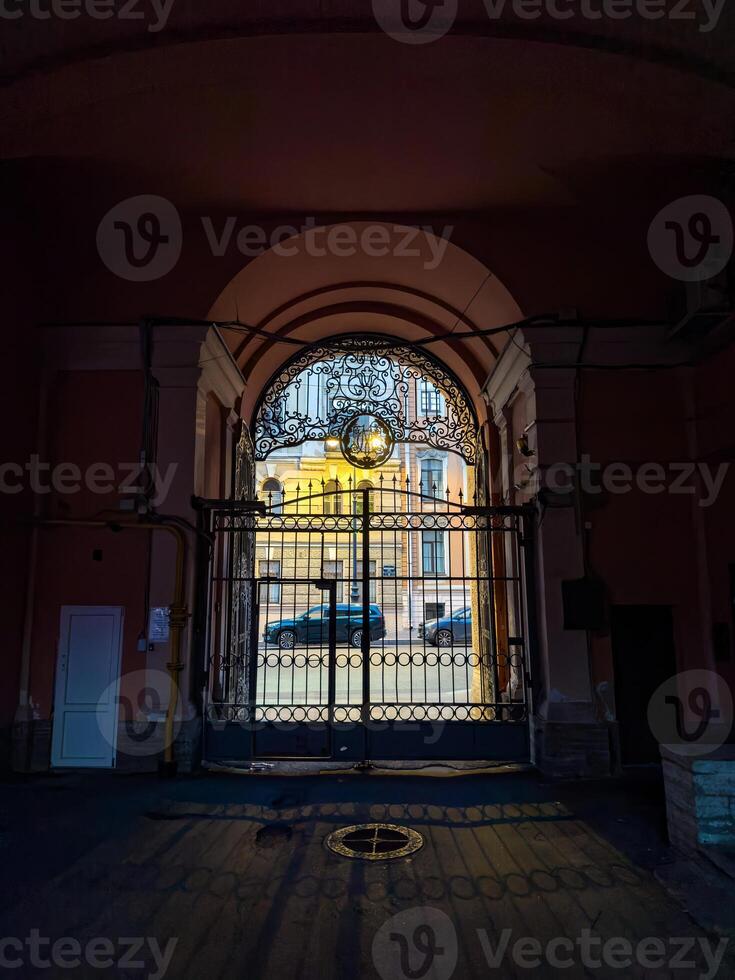 View through a gated archway showcasing intricate ironwork and a glimpse of a historic urban street bathed in evening light. Captured in a low-light setting, highlighting the contrast between the interior shadow and exterior illumination photo