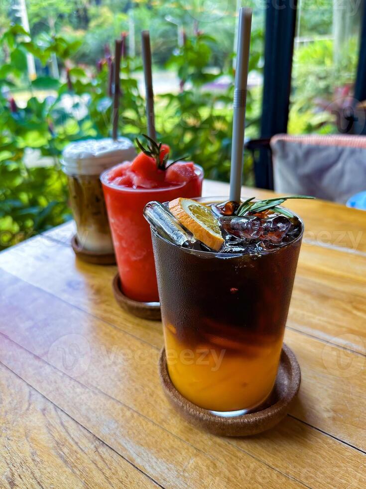 A selection of refreshing drinks including iced coffee, watermelon smoothie, and citrus iced tea on a wooden table, set against a lush tropical backdrop. Perfect for food and beverage themes. photo