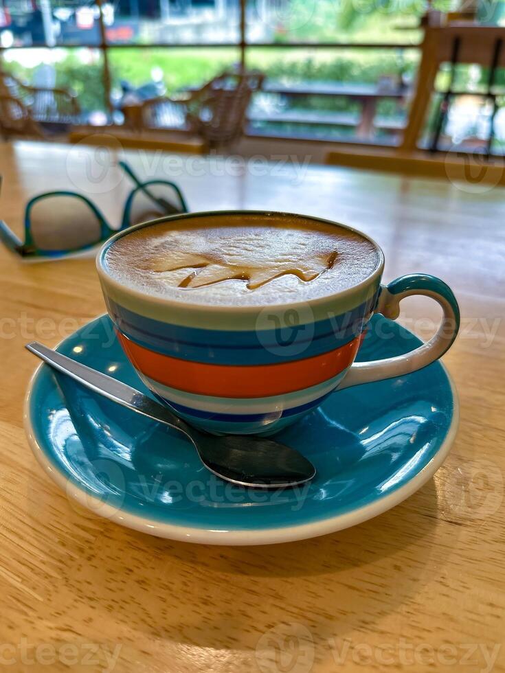A close-up of a creamy cappuccino served in a vibrant striped cup with a matching saucer and spoon, set on a wooden table in a cozy cafe, with sunglasses and a scenic view in the background photo