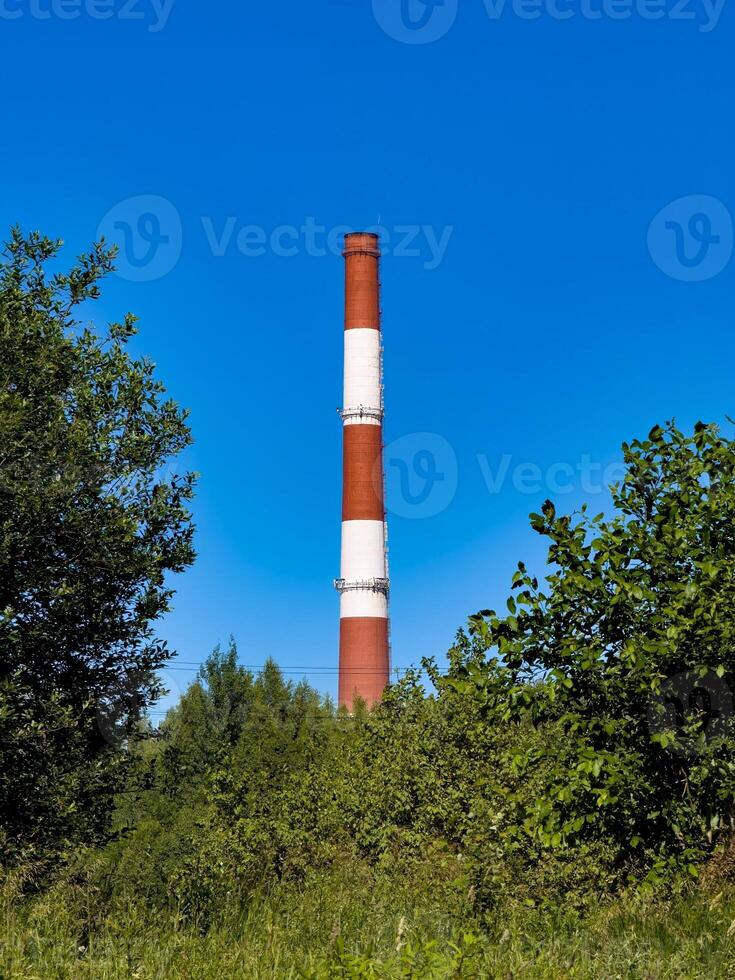 A towering industrial chimney rises above lush green foliage under a clear blue sky. This juxtaposition of nature and industry is ideal for environmental and industrial-themed projects photo