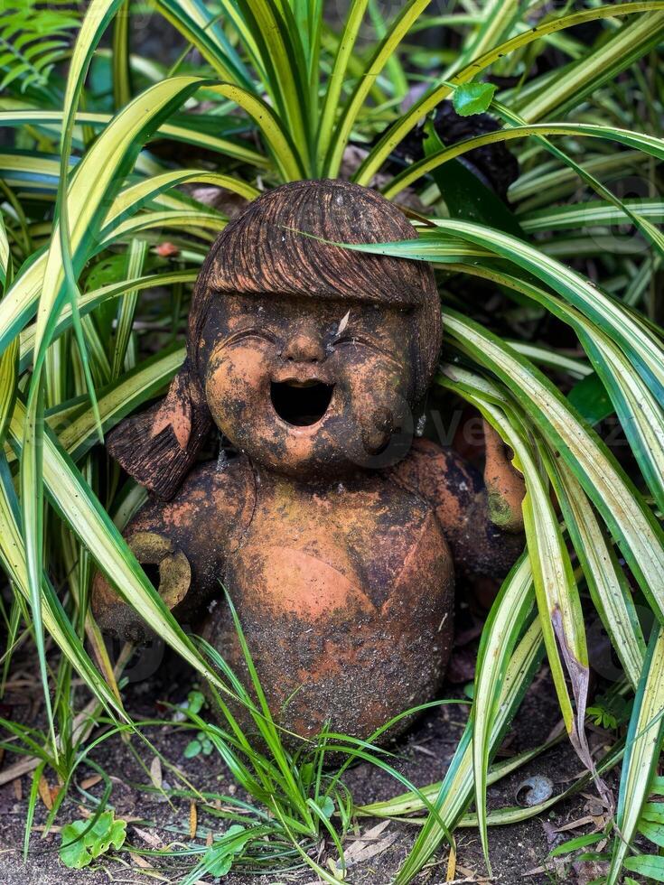 A rustic clay doll statue with a joyful expression, nestled amidst vibrant green foliage in a garden. Perfect for garden decor, nature, and artistic themes photo