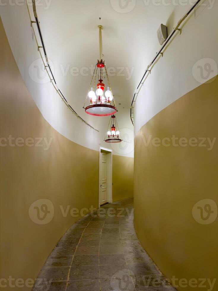 A narrow, curved hallway illuminated by vintage red chandeliers hanging from a vaulted ceiling. The hallway features beige walls and stone tiles, leading to a closed white door, creating a mysterious and historical ambiance photo