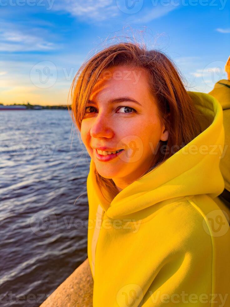 A close-up portrait of a young woman enjoying the sunset by the river in St. Petersburg, Russia. Her bright yellow hoodie glows warmly in the golden hour light, capturing a moment of joy and serenity photo