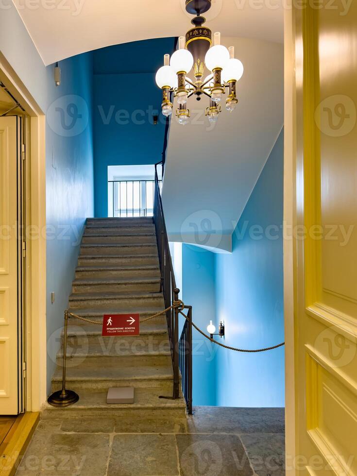 A vintage staircase illuminated by a classic chandelier in a historic building. The staircase features stone steps, ornate iron railings, and blue walls, creating an elegant and timeless interior space photo