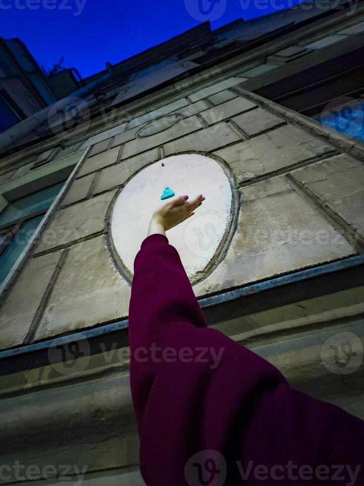 A cheerful woman in a maroon sweater reaches up towards a small blue Buddha figurine attached to the wall of a historic building. The playful interaction with the unique street art adds a touch of whimsy to the urban scene photo