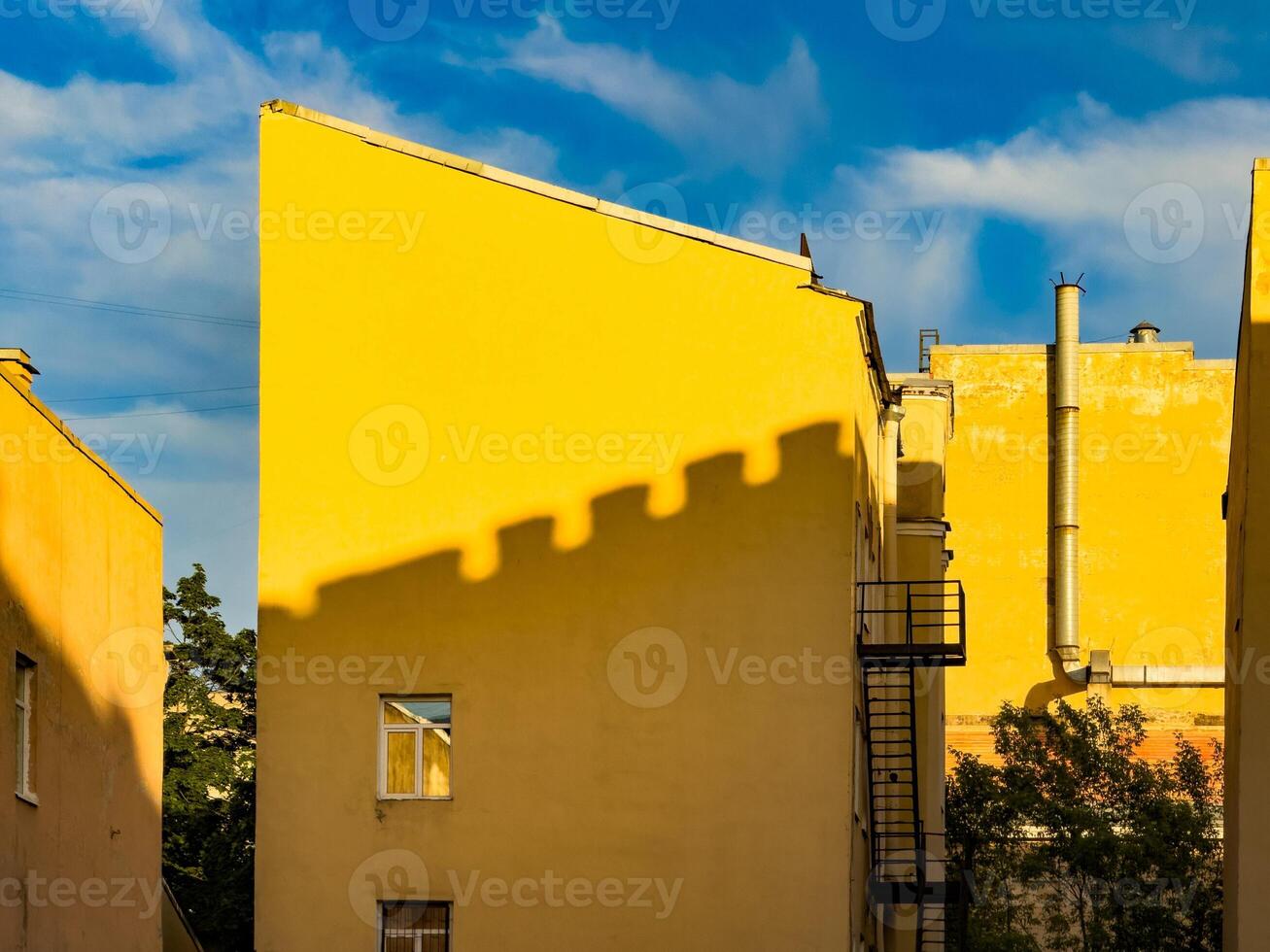 A striking image of bright yellow buildings in St. Petersburg, Russia, with shadows creating intriguing patterns on the walls. The vibrant yellow contrasts beautifully with the blue sky, creating a captivating and cheerful urban scene photo
