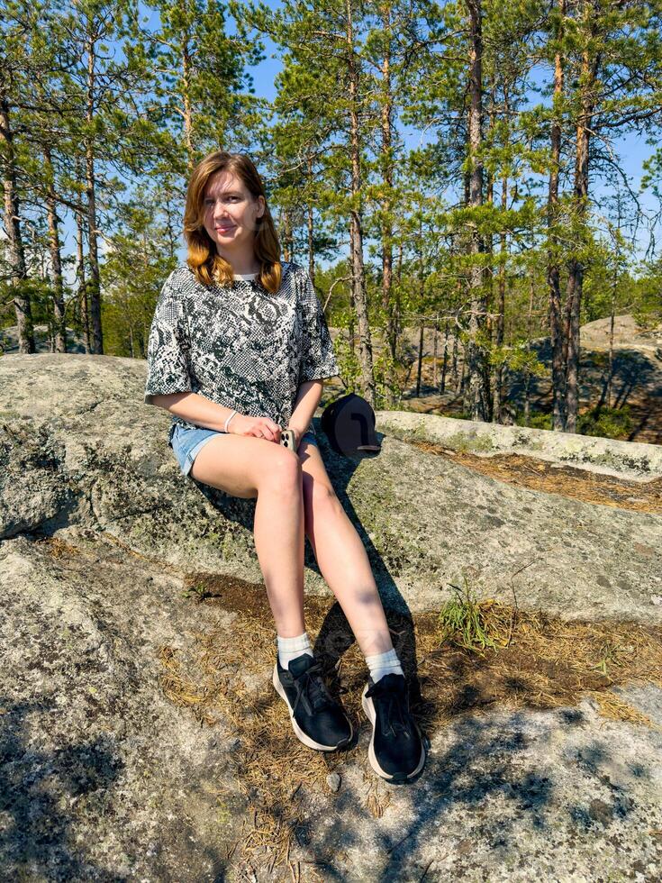 A young woman sitting on a rock in a pine forest under a clear blue sky, enjoying the serenity of nature. Perfect for themes of outdoor adventure, relaxation, and nature appreciation photo