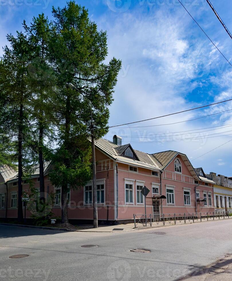 A picturesque pink wooden house sits gracefully at a sunny street corner, framed by tall trees and a clear blue sky, embodying a quaint and peaceful charm. photo