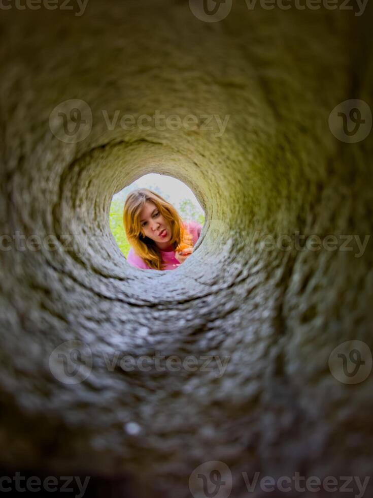 A young girl peers down a hole in granite, her face framed by the rugged stone. Her curious expression and the unique perspective create an engaging and playful scene, perfect for illustrating curiosity, exploration, and childhood adventures. photo
