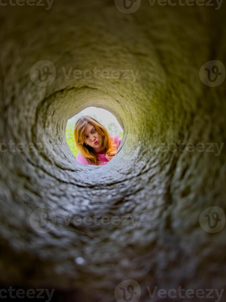 A young girl peers down a hole in granite, her face framed by the rugged stone. Her curious expression and the unique perspective create an engaging and playful scene, perfect for illustrating curiosity, exploration, and childhood adventures. photo