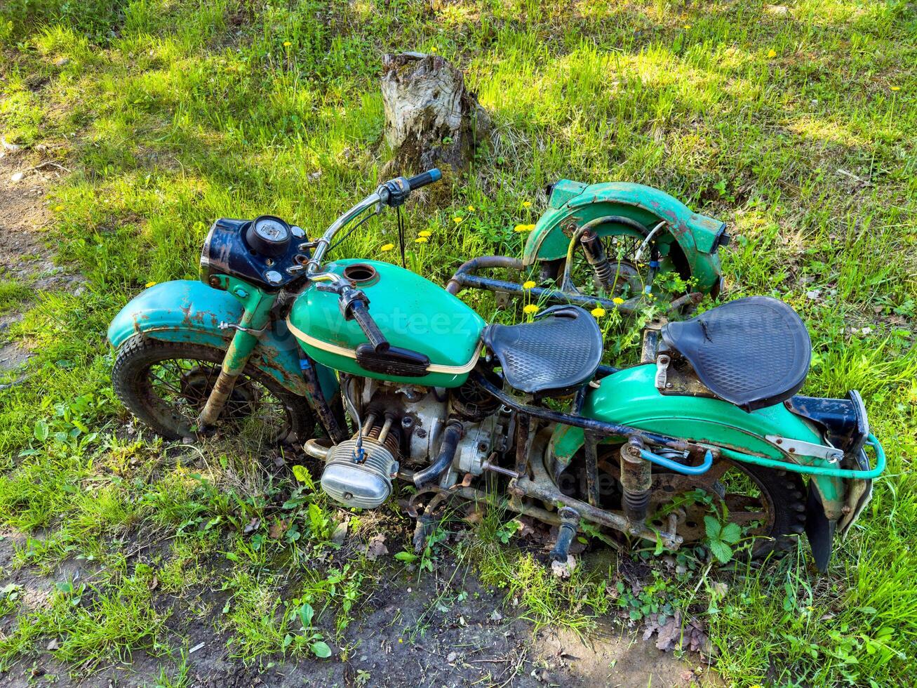 An old, abandoned green motorcycle sits partially buried in overgrown grass. The weathered, rusted parts and vibrant green surroundings create a nostalgic scene, perfect for themes of decay, history, and forgotten treasures. photo