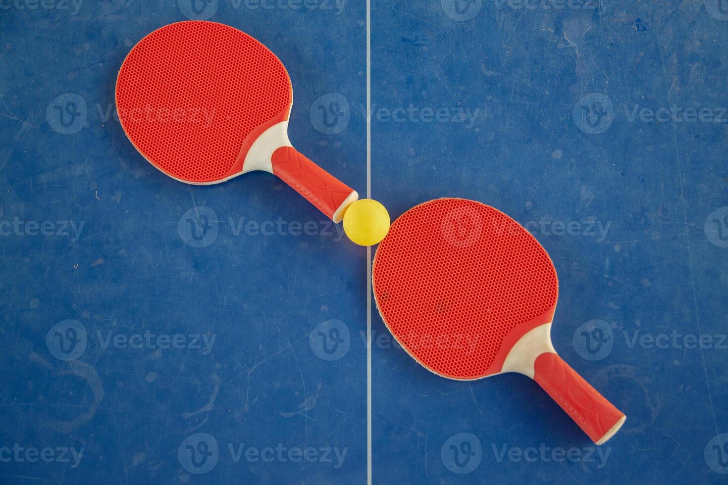 Red table tennis rackets with a yellow ball between them on a blue background, ping pong sports equipment, symmetry concept regarding the center of the table with a white line, top view, photo