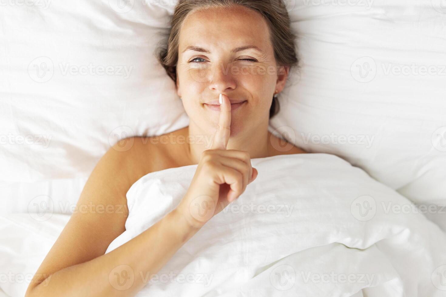 Portrait of a woman lying in bed covered with a blanket holding finger on lips photo