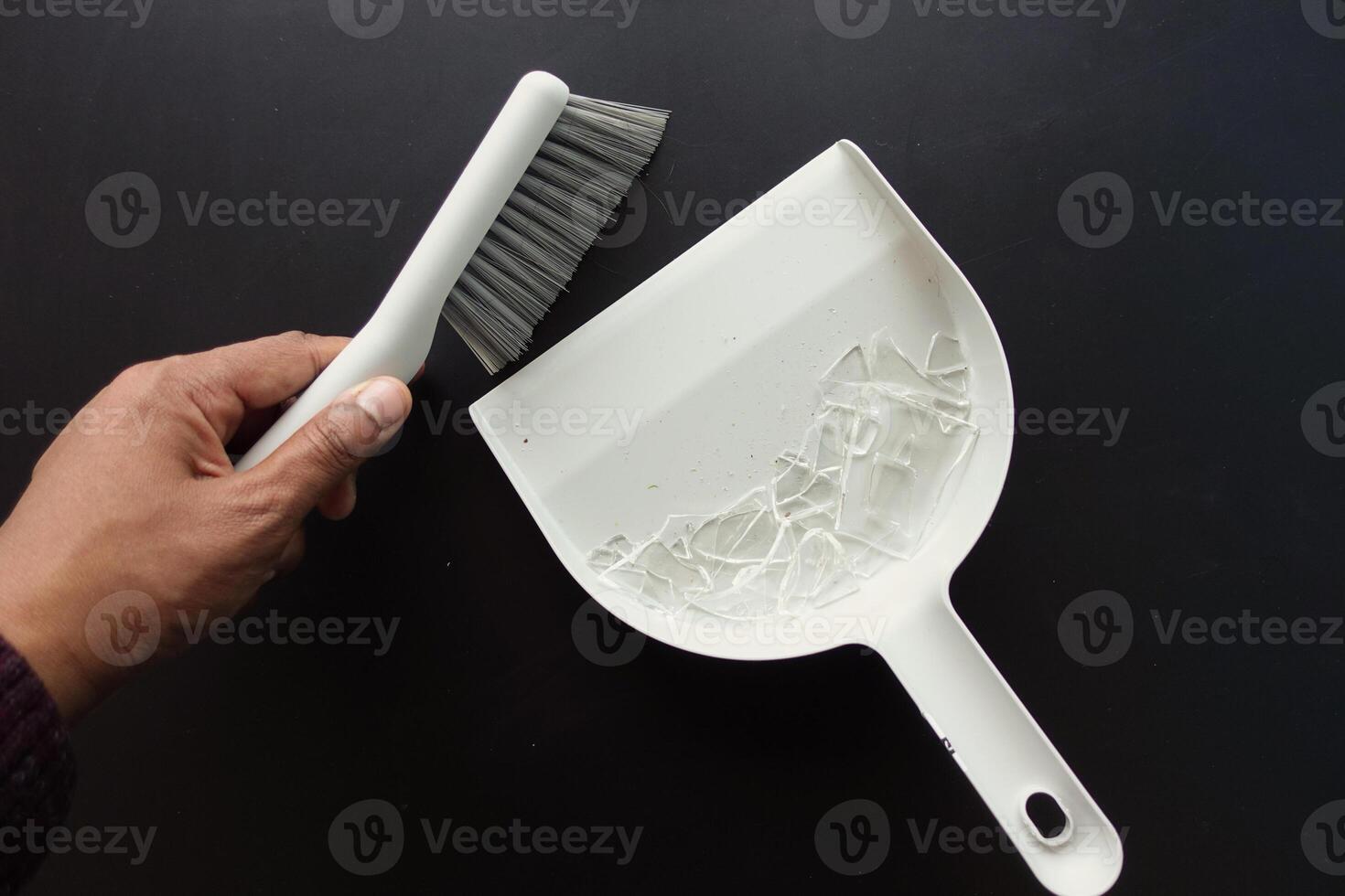 A hand holding a white dustpan and brush set for housekeeping against a dark background photo