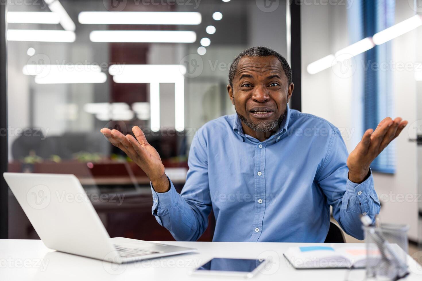 Businessman in blue shirt sitting at desk with laptop and tablet, gesturing confusion in modern office. Concept of frustration, problem-solving, misunderstanding, and communication issues workplace. photo