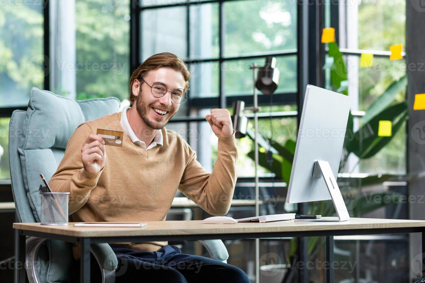 A young man successfully uses a credit card. He sits happily in the office at the computer, holds a card, looks at the camera, shows a yes gesture, celebrates. photo