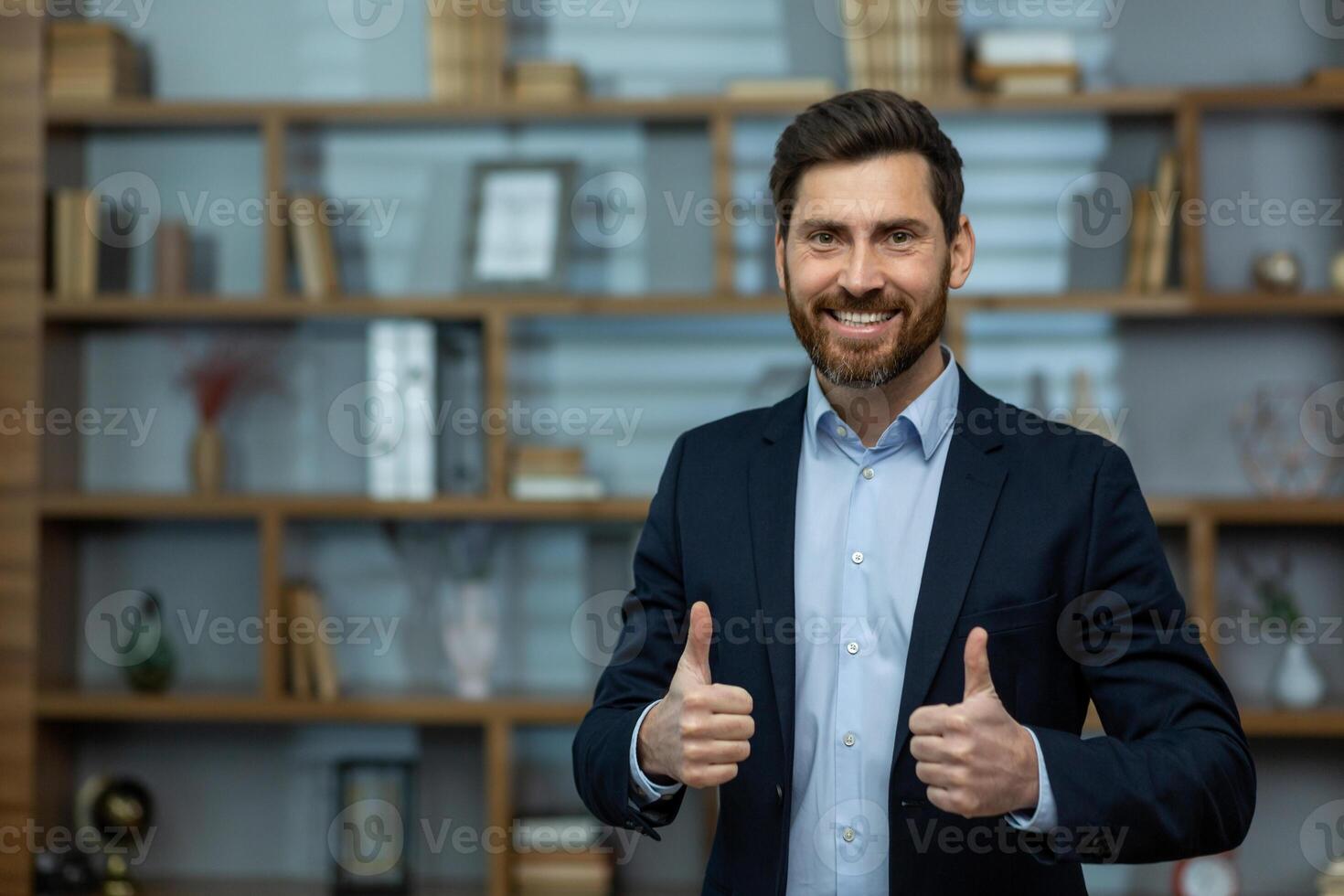 Caucasian businessman in suit giving thumbs up gesture while standing on background of office. Successful male making positive and approving sign with both hands while looking at camera indoors. photo