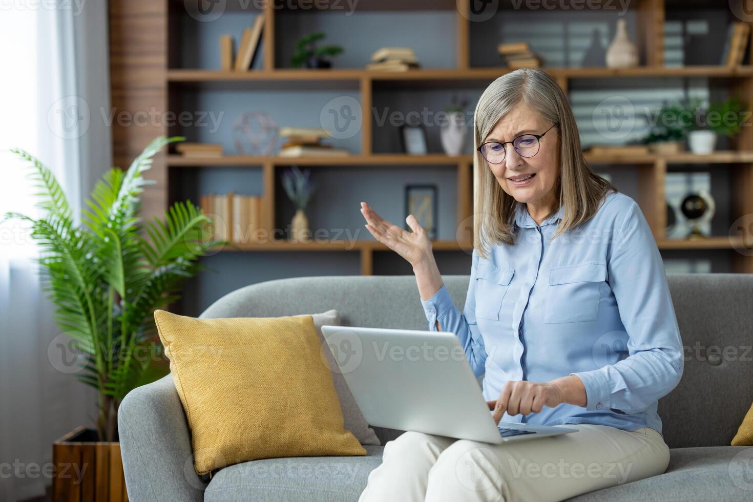 Senior woman sitting on a sofa, frustrated with computer problems while working from home. She looks confused and is gesturing with her hand, indicating technical issues with her laptop. photo