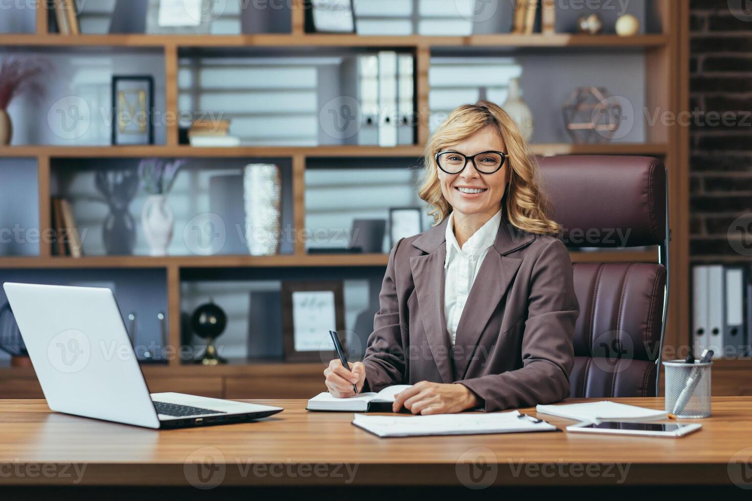 Smiling experienced blonde businesswoman working with laptop at a tidy office desk, exuding leadership and professionalism. photo