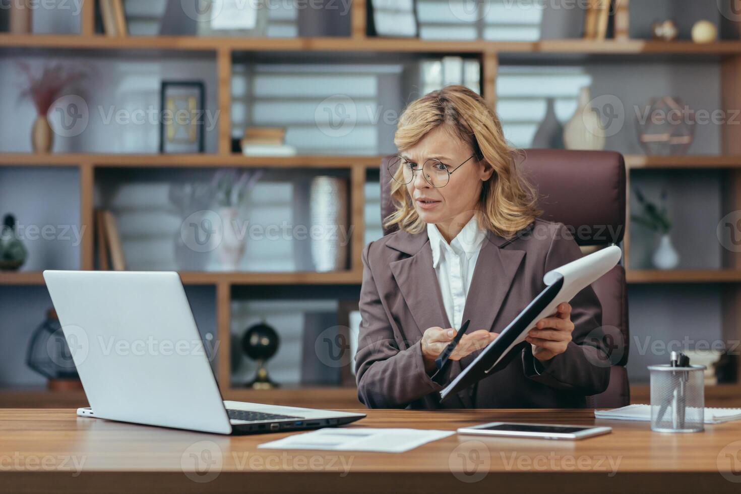 Online business meeting. A senior woman in a suit is sitting in the office at a desk and working on a laptop, talking on a call and anxiously showing documents to the camera. photo