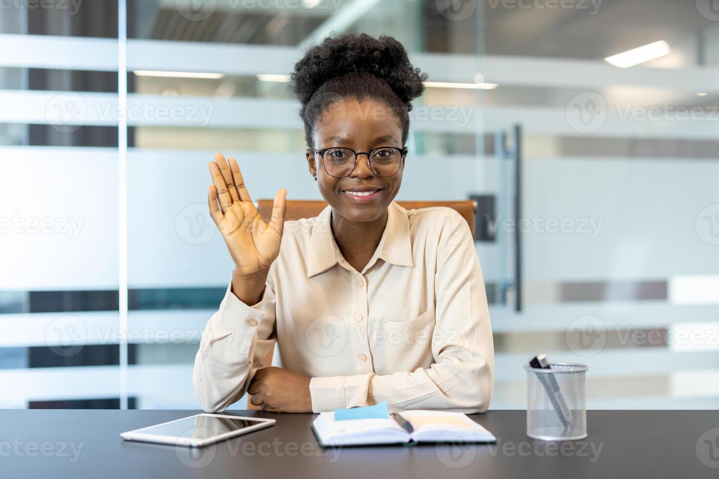Businesswoman waving hello during a call in modern office setting. Notebook, tablet, and pen holder on desk. Professional and friendly working atmosphere. photo