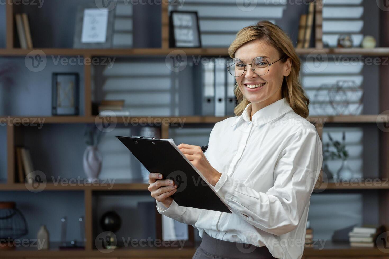 Portrait of an older female teacher, tutor, lecturer in glasses and a white shirt. He stands in the office, library with a folder and a pen in his hands, writes, looks at the camera, smiles. photo