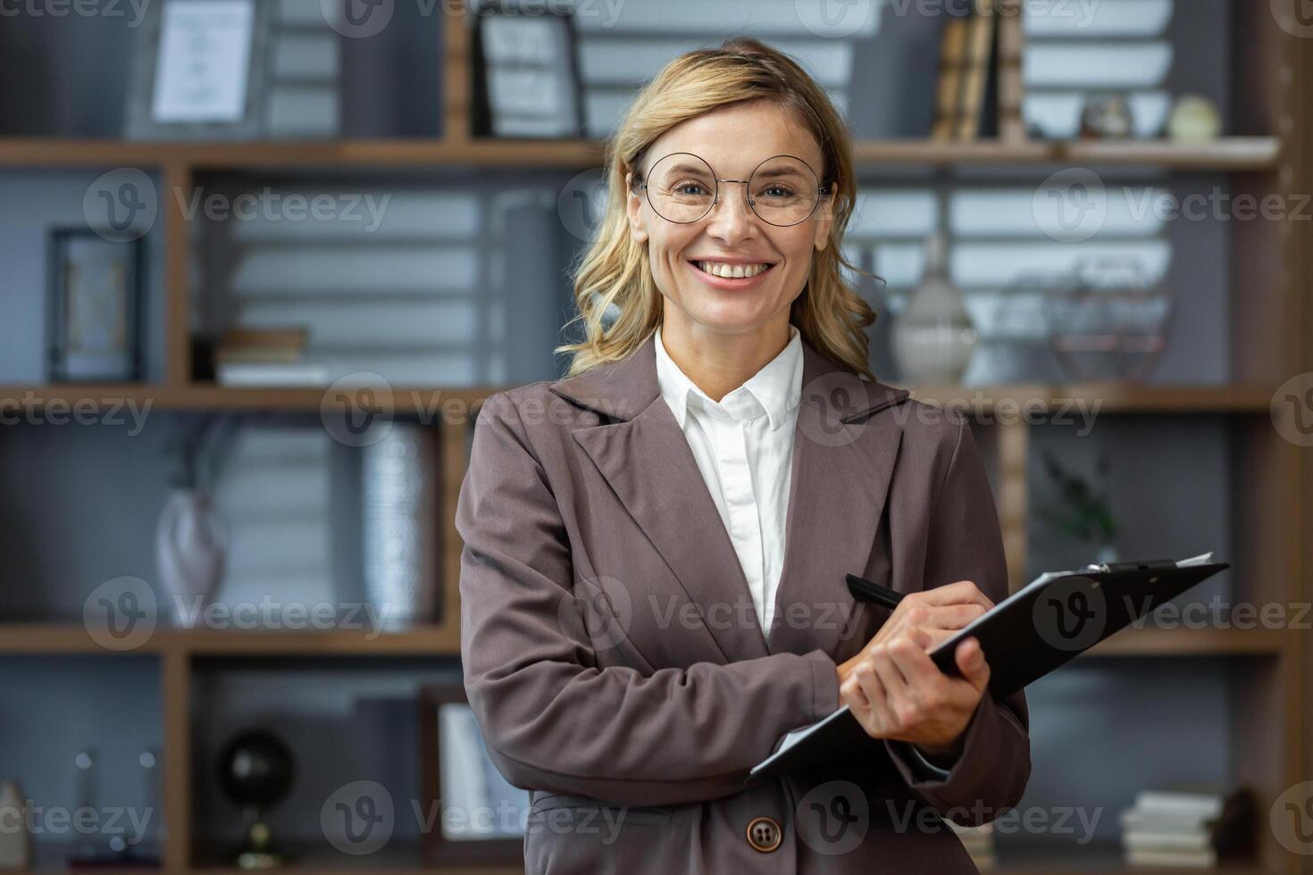 Portrait of an older female teacher, tutor, lecturer in glasses and a suit. Standing in the office with a folder and a pen in his hands, looking at the camera, smiling. photo