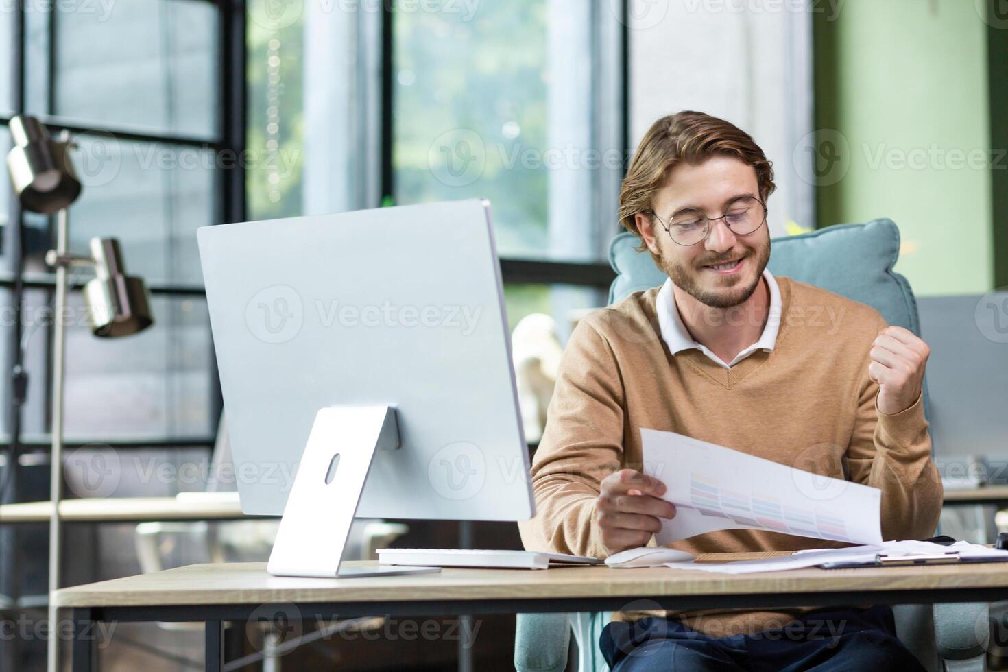 A happy manager in the office rejoices at the concluded contract, holds documents in his hands, joyfully raises his hand, happiness at the completion of work. photo