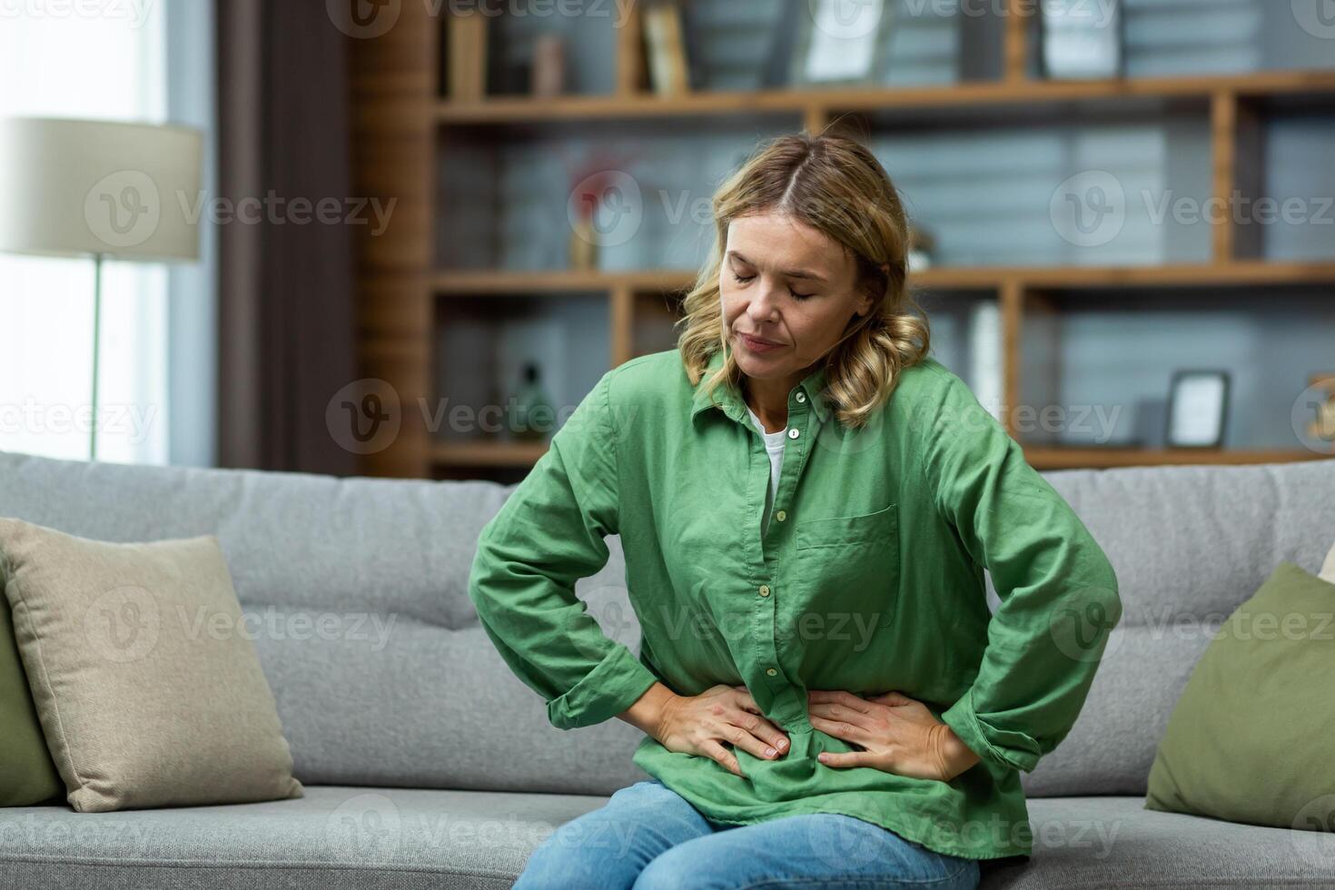 An elderly woman is sitting on the sofa at home, holding her stomach with her hands. Feels the pain of internal organs, indigestion, poisoning, menstrual pain. photo