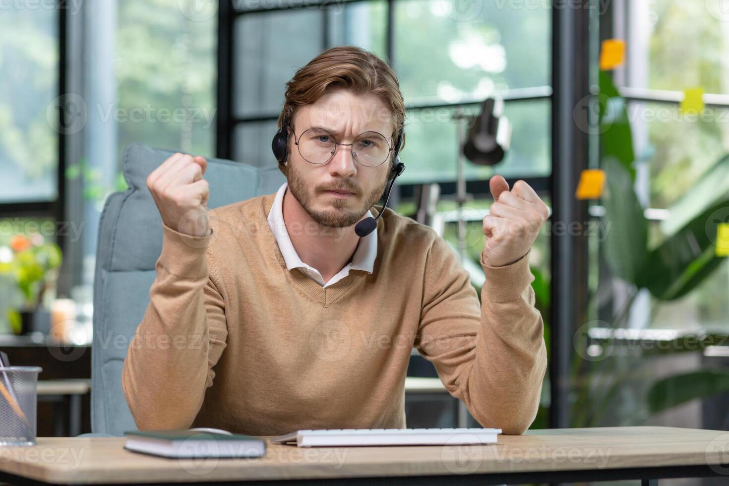 An angry young man, a businessman, a programmer in a headset, sits in the office and speaks expressively to the camera, holds his fists in his hands. Online meeting, discussion, deadline, project. photo