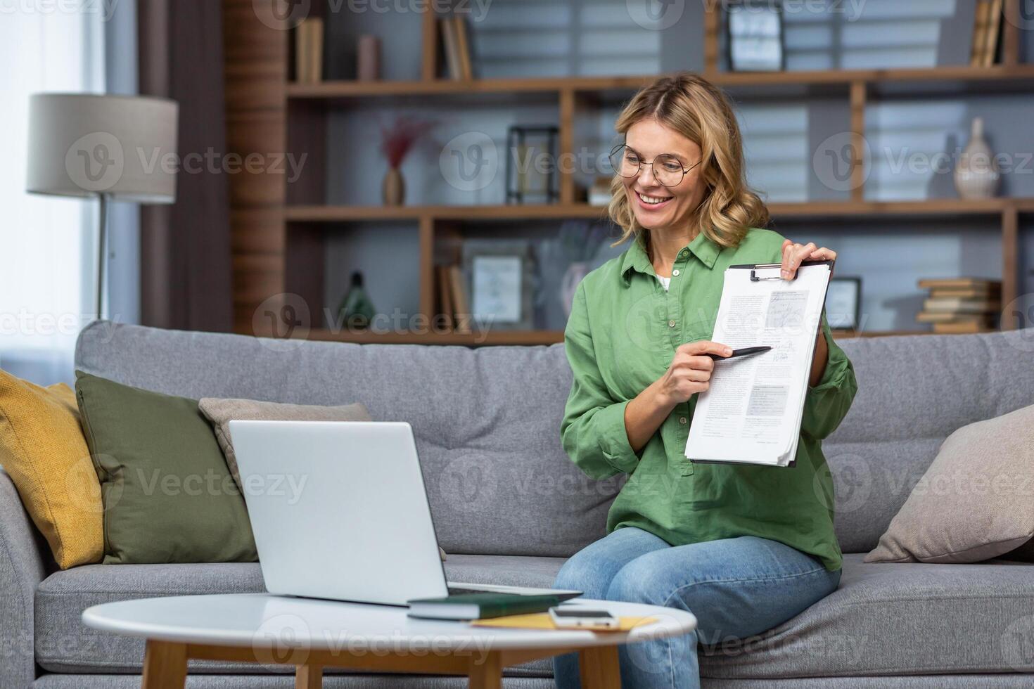 Portrait of a woman at home teaching online remotely, the teacher sits on the sofa and shows a document to the camera of the laptop online classes. photo