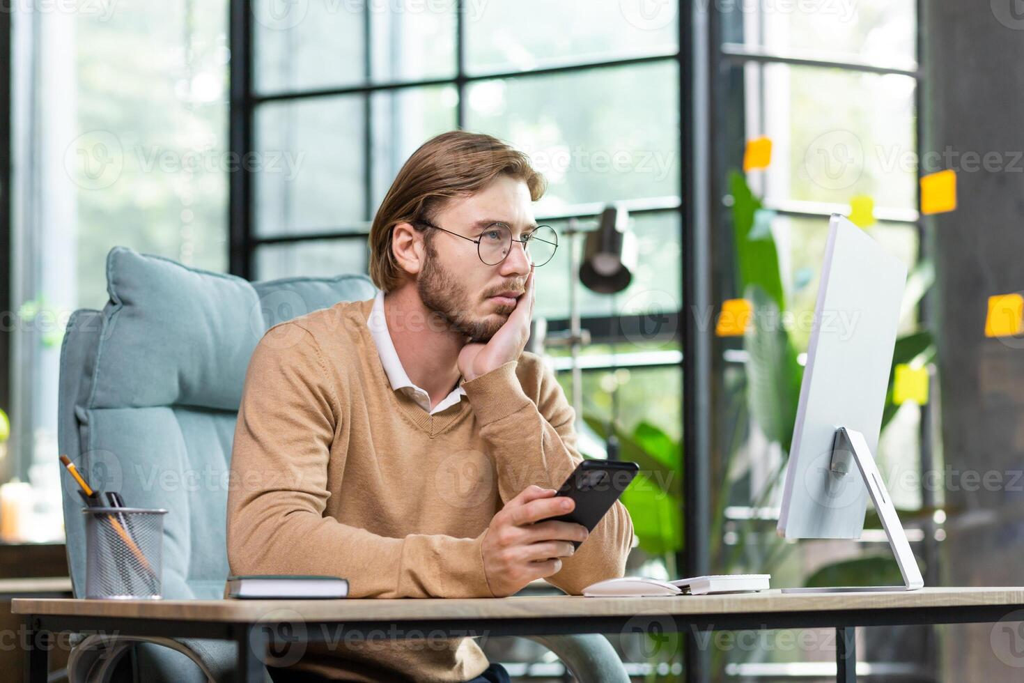 Upset man sitting at the table with a phone in his hands looks tiredly at the computer screen, holds his head with his hand, overworked burnout. photo